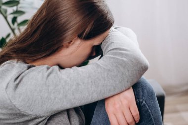 Mutsuz, kaygılı, yüzünü diziyle kapatan genç bir kadın. Oturma odasında kanepede oturuyor. Kalbi kırık, depresyondaki bir kadın.