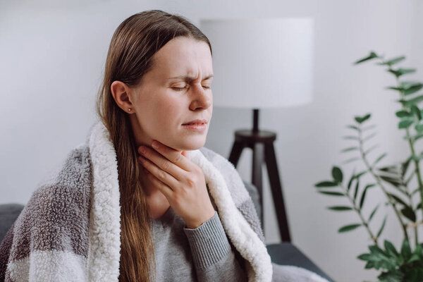 Symptoms from flu season in winter concept. Close-up of unhealthy sad young woman covered blanket sitting on grey couch at home suffering from sore throat, angina, hard to swallow, voice loss