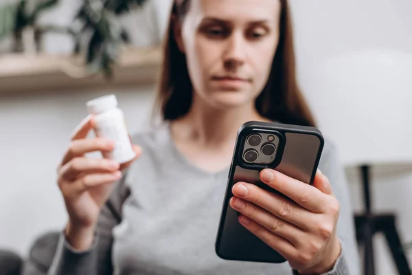 Selective focus of young lady using smartphone for reading, searching prescription on bottle medicine, pill label text about information online, instructions side effects, pharmacy medicament concept
