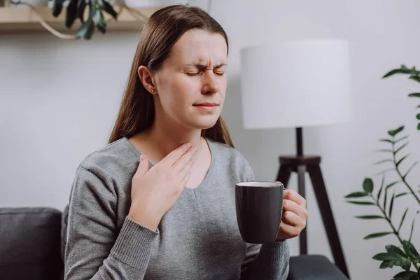 stock image Sick unhappy young caucasian female 25s touch neck, have sore throat, holding mug, drink warm water, have a fever, flu in weakness, sitting alone on couch at home. Health care on virus seasonal