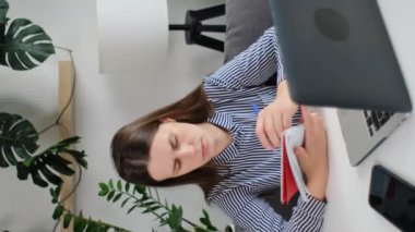Dikey videoda, esmer bir kız dizüstü bilgisayarda internet platformunda oturup internetten ders çalışıyor, notlar yazıyor. Genç serbest çalışan kadın iş projesinde çalışıyor.