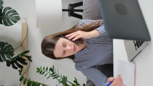 かわいい若い女性の垂直ビデオは ペンを詰め込む情報を保持し ノートを取る 携帯電話の仕事について話しながらノートパソコンで座って 顧客サービスマネージャーとの正式な会話を与える — ストック動画