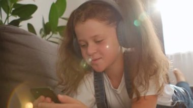 Kulaklık takmış, elinde telefonla koltukta uzanan mutlu şirin kız çocuğu. Akıllı telefon teknolojisi kullanan küçük bir çocuk evdeki oturma odasında cep telefonuyla internetten video dersleri alıyor.