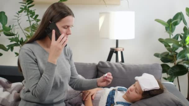 悲伤的十多岁的女儿躺在沙发上 觉得发烧得了感冒 焦急的母亲在测量儿童体温后 把电话举到耳边给家庭医生打电话 叫家庭医生 — 图库视频影像