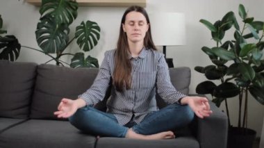25 'li yaşlarındaki sakin genç bir kadın nilüfer pozisyonunda gözleri kapalı ve parmakları katlanmış bir şekilde evdeki rahat koltukta yoga antrenmanı yapıyor, nefes egzersizleri yapıyor stressiz tek başına vakit geçiriyor.
