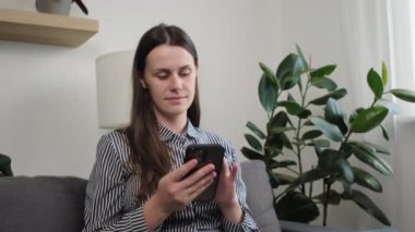 Kanepede oturan beyaz bir kadın akıllı telefon kullanıyor, mesajlarını paylaşıyor, sosyal medyada erkek arkadaşıyla sohbet ediyor ve evde boş vakitlerin tadını çıkarıyor. Modern kablosuz teknoloji kullanımı, sanal iletişim kavramı