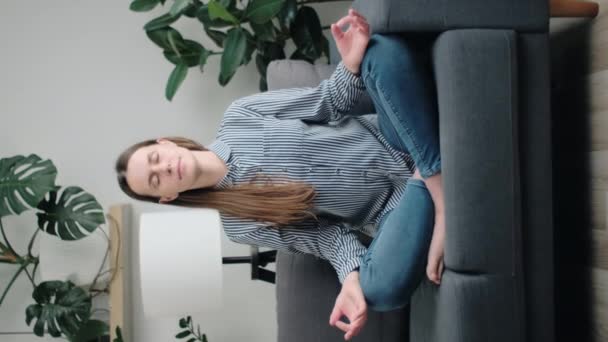 年轻可爱女子交叉腿坐在沙发上的垂直镜头 做冥想练习 促进内部平衡 改善自我意识 思维状态 和谐和呼吸练习 — 图库视频影像