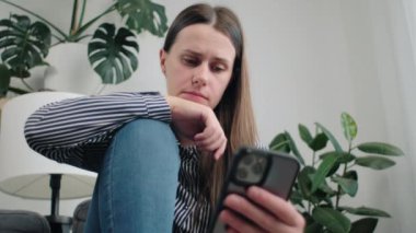 Düşünceli, genç, beyaz bir kadın evdeki koltukta oturmuş, cep telefonuyla sorunu düşünüp duruyor. Endişeli üzgün esmer kız 25 'li yaşlarda akıllı telefondaki mesaj ya da mesajdan rahatsız olur