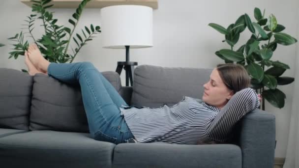 可爱的梦幻般的年轻的高加索女人躺在舒适的沙发上 躺在家里的客厅里 双腿靠着背 望着别处 享受家庭休闲和康复的理念 — 图库视频影像