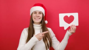 Neşeli genç kadın sıcak beyaz süveter giyer ve Noel Baba şapkası elinde simge şeklinde bir kalp taşır. Pastel kırmızı stüdyo arka plan duvarında izole bir poz verir. Mutlu yıllar 2024 tatil konsepti