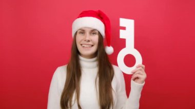 Mutlu, güzel genç bir kadın rahat bir süveter ve Xmas Noel Baba şapkası giyer. Elinde büyük beyaz bir anahtar vardır. Stüdyodaki kırmızı renk arka plan duvarında izole bir şekilde poz verir. Mutlu yıllar 2024 bayram konsepti.