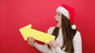 Mutlu genç kadın, kırmızı stüdyo arka plan duvarında izole edilmiş oklu reklam için fotokopi alanını gösteren Noel şapkası ve beyaz süveter giyer. Mutlu yeni yıl kutlamaları, mutlu tatiller.