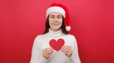 Mutlu, sevimli, genç, canlı, 25 'lik Noel Baba. Beyaz, sıcak kazak ve küçük kırmızı bir kalp taşıyan Noel şapkalı. Kameraya mutlu bir şekilde bakıyor, stüdyodaki pastel kırmızı arka plan duvarında izole edilmiş poz veriyor.