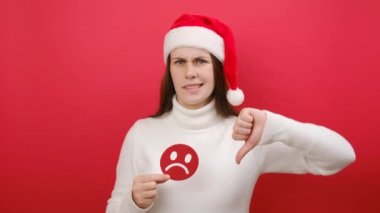 Kızgın genç kadın beyaz süveter ve Noel Baba şapkası takıyor, elinde tatminsiz üzgün surat ifadesi var, baş parmağını aşağı gösteriyor, stüdyoda kırmızı arka planda izole edilmiş poz veriyor. Yeni Yıl kavramı, olumsuz duygu