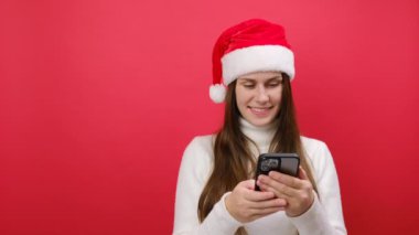 25 'li yaşlarındaki neşeli genç kadın beyaz süveter ve Xmas Noel Baba şapkası takıyor cep telefonu kullanıyor ve Pastel Red stüdyo arka planında izole bir şekilde poz veriyor. Mutlu yıllar 2024 bayram konsepti.