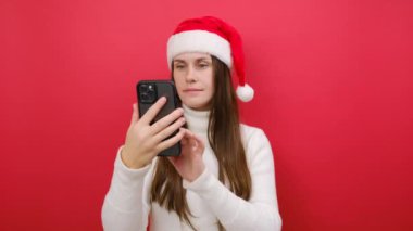 Samimi beyaz kazak giyen mutlu genç bir kadın ve cep telefonunda Xmas Noel Baba şapkası parmağıyla stüdyodaki pastel kırmızı arka plan duvarında izole bir şekilde poz veriyor. Mutlu yıllar 2024 tatil konsepti