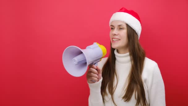 かなり若い女性は白いセーターを着用し メガフォンでXmas Santaハットの叫び声は販売を発表し スタジオでパステルレッドカラーの背景の上に隔離されています ハッピーニューイヤー2024のお祝いの休日のコンセプト — ストック動画