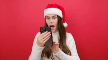 Neşeli genç bir kadın rahat bir süveter giyer ve Noel Baba şapkası takar cep telefonu kullanır kazanan hareketi yapar, pastel kırmızısı stüdyo arka planında izole edilmiş poz verir. Mutlu yıllar 2024 tatil konsepti