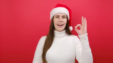 Mutlu, güler yüzlü genç bir kadın sıcak beyaz süveter ve Noel Baba şapkası giyer. Tamam jest, stüdyodaki pastel kırmızı arka plan duvarına izole edilmiş poz verir. Mutlu yıllar Noel 2024 konsepti