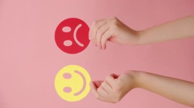 Kadının elinde pembe duvarda izole edilmiş mutlu ve kızgın bir yüz kağıdı olan dikey bir video. Geri bildirim derecesi, duygusal zeka, duygu kontrolü, zihinsel sağlık değerlendirmesi, bipolar bozukluk konsepti