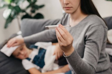 Beyaz tenli genç annenin seçici odak noktası. Kızının vücut ısısını ölçüyor. Kafa, alın ve termometreyi tutuyor. Hasta küçük kız kanepede yatıyor, gripten muzdarip, ateşi var.