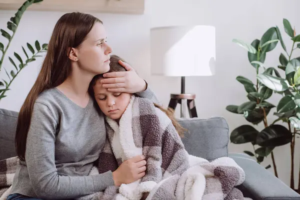 Caring young mother hugging, sick upset little daughter covered blanket, sitting on sofa at home, worried mum comforting sad ill kid girl, expressing love and support, child psychologist concept