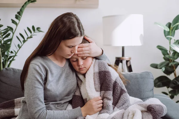 Loving young mother hugging sick upset little daughter covered blanket, sitting together on couch at home, caring mum comforting sad ill kid, expressing love and support, child psychologist concept