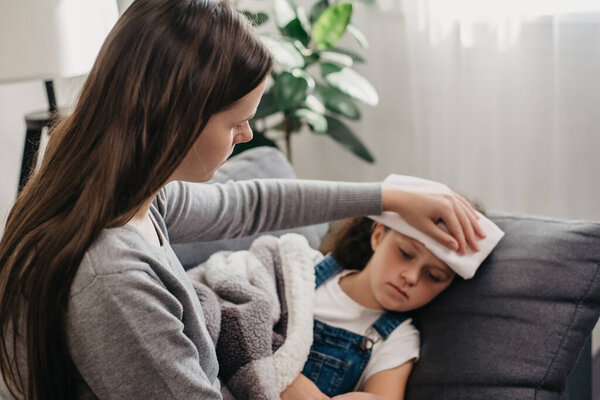 Выборочный фокус заботливой молодой матери положил полотенце на голову больной девочки с высокой температурой тела лежал на диване дома. Мамочка заботится о больной маленькой дочери. Уход за детьми, концепция детского здравоохранения