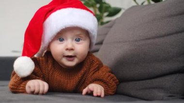 Neşeli, tapılası bebek portresi, kameraya bakmaktan mutlu, Noel şapkası takan bebek ve evdeki rahat kanepede uzanan sıcak örülü süveter. Xmas tatil konsepti
