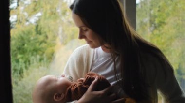 Mutlu, sevgi dolu bir anne, güzel bir bebek, sakin bir sonbahar arka planında, pencerenin kenarında oturuyor. Sevecen, güler yüzlü bir anne şımarık küçük bir bebeği evde okşuyor. Annelik kavramı