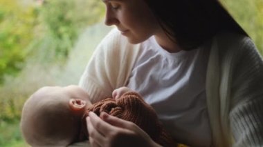 Sevgi dolu bir anne, sevimli bir bebek, sakin bir sonbahar doğası arka planında, rahat bir pencere kenarında oturuyor, sıcak öpücükle tatlı bir çocukla konuşuyor. Şefkatli anne şımarık küçük bebek çocuğu okşuyor.