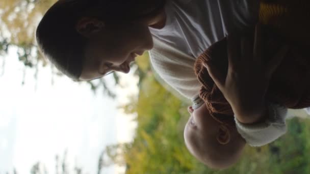 美しい秋の自然の背景に居心地の良い窓に座って愛らしい赤ん坊の息子を抱く幸せな世話をする若い白人ママの垂直ショット かわいい 小さな ベイビー — ストック動画