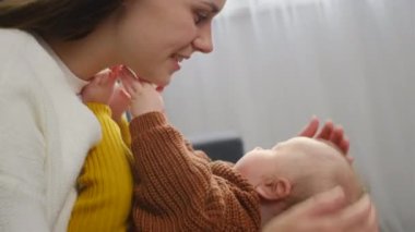 Mutlu, genç, sevgi dolu anne rahat koltukta oturan küçük ayakları öpüyor, yeni doğmuş bebek ayak parmakları kalın kazak giyiyor. Çocukla, anneyle ve çocukla duygusal anlar, anne bakımı konsepti.