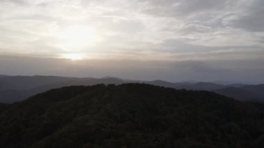 Bulutlu gökyüzünün ve ağaçların sinematik drone görüntüleri günbatımında ya da gündoğumunda sonbahar tepelerini kaplar. Sonbahar akşamları dağ ormanı manzaralı hava uçuşları. Yaprak döken ve yemyeşil ağaçlar