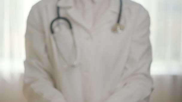 診療所に立っている手の小さな紙の家で握る専門の医学の白い制服を身に着けている認識できない若い女性の医師の近く 医療生活保険コンセプト — ストック動画
