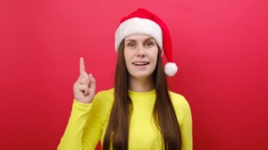 Sevimli genç bir kadın sıcak sarı süveter ve Noel Baba şapkası giyer. Kırmızı stüdyo arka planında izole edilmiş harika bir fikirle işaret parmağı tutar. Mutlu yıllar 2024 Noel tatili konsepti