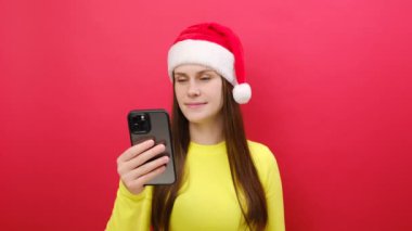 Mutlu genç kadın sarı süveter ve Noel Baba şapkası takıyor cep telefonu kalp şeklini ikon işareti gibi kullanıyor ve pastel kırmızısı arka planda izole edilmiş poz veriyor. Mutlu yıllar 2024 tatil konsepti