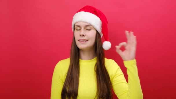 メリーは黄色いセーターとサンタクロースの帽子をかぶった25代の笑顔の若い女性に満足し スタジオのパステル赤い背景の壁の上に隔離された良いジェスチャーを見せます ハッピーニューイヤークリスマス2024コンセプト — ストック動画