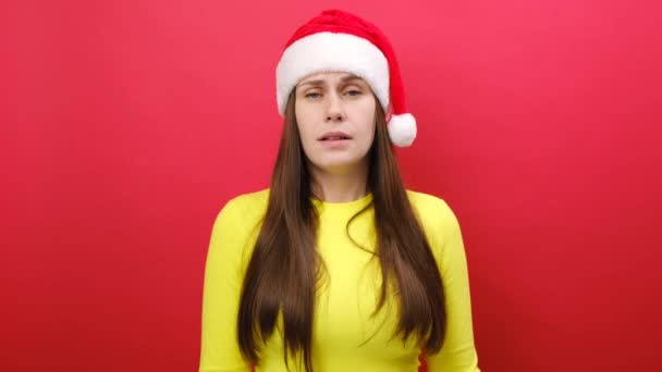 年轻的女人穿着黄色的毛衣 头戴圣诞帽 耸了耸肩 毫无头绪地展开双手 摆出孤立的姿势 笼罩在苍白的红色画室的背景上 新年快乐2024假期的概念 — 图库视频影像