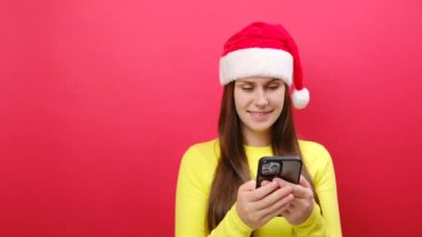 Mutlu rüya gören genç kadın sarı kazak giyiyor ve Noel Baba şapkası takıyor cep telefonu kullanıyor. Bölgeye bak, pastel kırmızı arka planda izole edilmiş poz veriyor. Mutlu yıllar 2024 tatil konsepti