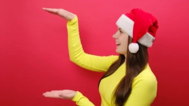 Neşeli genç kadın sarı süveter ve Noel Baba şapkası giyiyor. Çalışma alanının büyüklüğünü gösteren hareketler yapıyor. Kırmızı stüdyo arka plan duvarında izole edilmiş. Mutlu yıllar 2024 bayram konsepti.