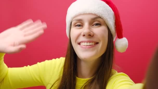 暖かい黄色いセーターを着て陽気な若い女性とサンタクロースは 赤いスタジオの背景の上に隔離された手を振って セルフィーをすることを嫌う ハッピーニューイヤー2024クリスマス休日のコンセプト — ストック動画
