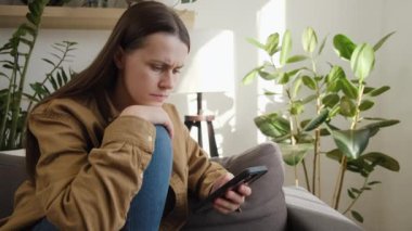 Ciddi üzgün, düşünceli, genç, beyaz kadın 25 'ler ellerinde akıllı telefon cihazıyla kanepede oturuyorlar. SMS' leri okuyorlar, üzülüyorlar, hoş olmayan haberler alıyorlar, internetten bilgi aldıklarını düşünüyorlar.