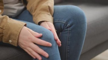 Evdeki kanepede otururken diz kaslarına masaj yapan kadın ellerini kapat. Bilinmeyen genç bir kadın diz eklem ağrısı çekiyor, yürüyemiyor, hastalık, kramp, romatizma konsepti...
