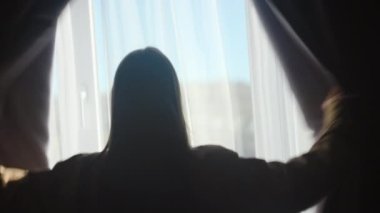 Siluet görünümlü beyaz kadın perdeyi açıyor ve pencereden bakıyor. Kendine güvenen milenyum hanımı sakin manzarayı izlemekten zevk alıyor, evde aktif bir yaz haftasonunun tadını çıkarıyor.