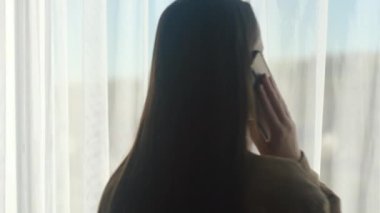 Evinin penceresinin yanında duran mutlu beyaz kadın silueti cep telefonuyla konuşuyor. Nesil ve modern teknoloji kullanımı, telefon görüşmesi etkinliği, düzen, iletişim konsepti