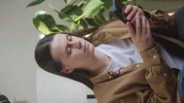 Mutlu bir milenyum kadınının kanepeye uzanmış, akıllı telefonu elinde tutarken dikey çekimi. Gülümseyen genç bayan sosyal ağlarda sohbet ediyor, komik videolar izliyor, evde mobil uygulamalar kullanıyor.