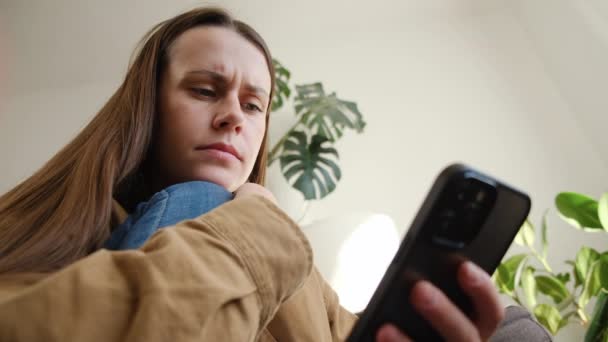 忧心忡忡的年轻女士用手机 得到了坏消息 看着别处 认真体贴的女人拿着智能手机 坐在沙发上 思考在线聊天短信 电子邮件 — 图库视频影像