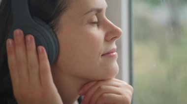 Sonbahar ormanı arka planında pencere kenarında oturan, kablosuz kulaklıklarla sakin klasik müzik dinleyen, hafta sonları evde tek başına tembel hobi yapan mutlu genç kadının yakın çekimi.
