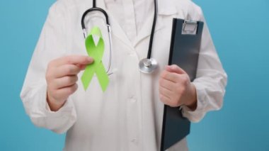 Mavi duvarda yeşil kurdele tutan üniformalı ve steteskoplu genç kadın doktorları yakından çek. Dünya bipolar günü. Akıl sağlığı farkındalığı günü, depresyon, beyin felci. Böbrek Günü konsepti